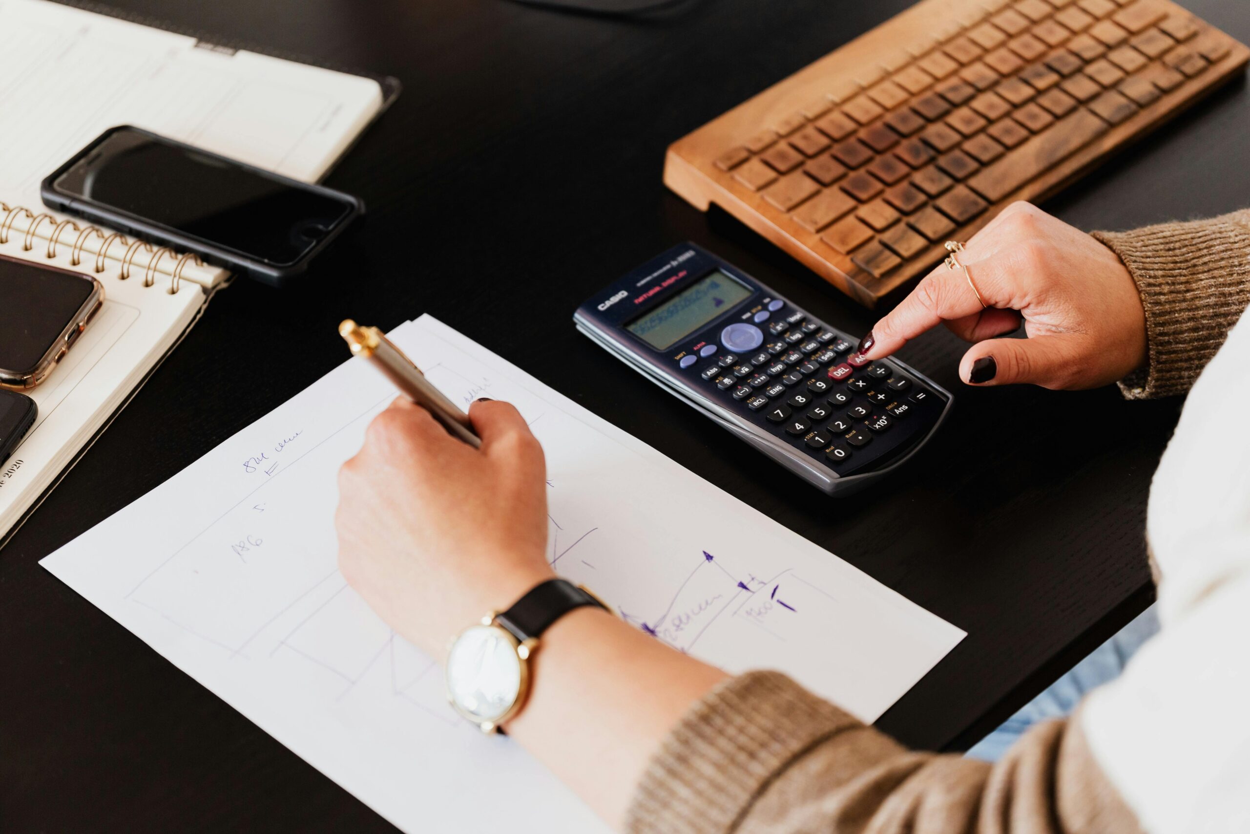 Close-up of hands working with a calculator and notebook on a desk, analyzing documents.