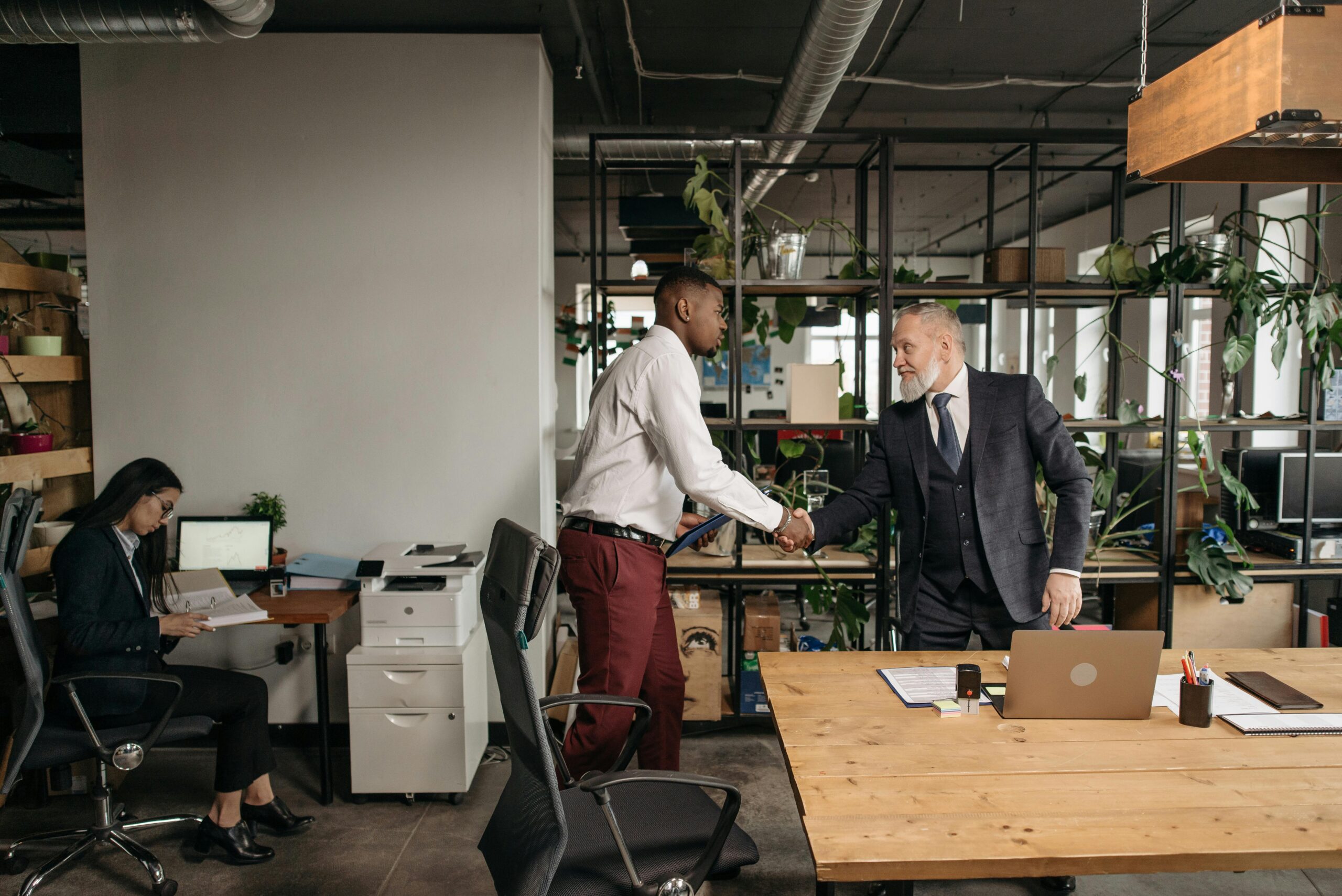 Business professionals exchanging a handshake in a modern office with coworker in the background.